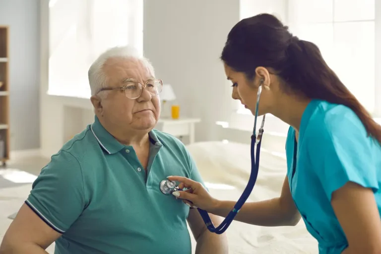 A female healthcare professional in blue scrubs leans forward to listen to the chest of an elderly man using a stethoscope